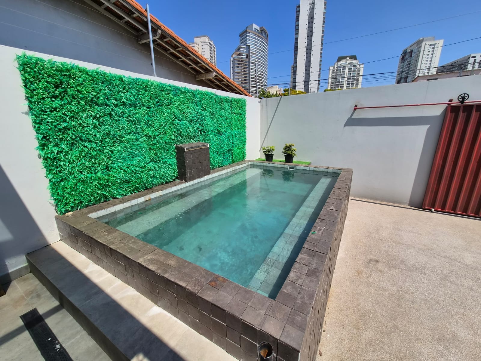 Piscina revestida com pastilhas, parede verde ao fundo e prédios de São Paulo no horizonte