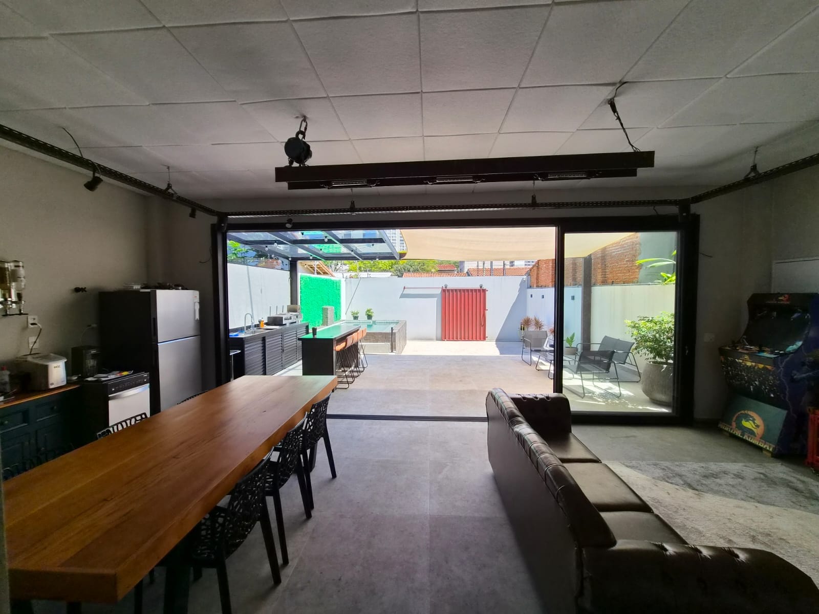 Sala integrada com mesa de madeira maciça, sofá de couro e portas de vidro totalmente abertas para a área gourmet e a piscina